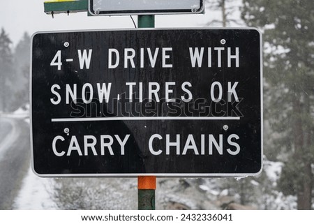 Highway sign in Sierra Nevada mountains that reads,"4-W Drive with Snow Tires OK -- Carry Chains"  Snowy winter backdrop with wet road.  Captured during a winter storm.  