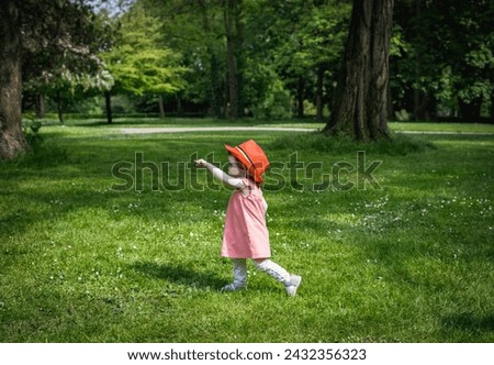 A portrait of one beautiful Caucasian little baby girl wearing a red Belgian flag hat stands sideways on the lawn in a city park on a summer day and points her finger to the left, close-up side view. Royalty-Free Stock Photo #2432356323