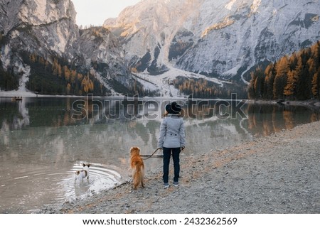 A person and their golden companion pause on a pebbled lakeshore, gazing at the serene alpine waters reflecting the glow of an autumn dusk Royalty-Free Stock Photo #2432362569