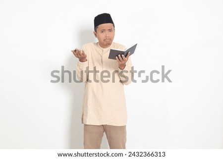 Portrait of confused Asian muslim man in koko shirt with peci difficulty understanding the contents of the book, reading a textbook. Isolated image on white background Royalty-Free Stock Photo #2432366313