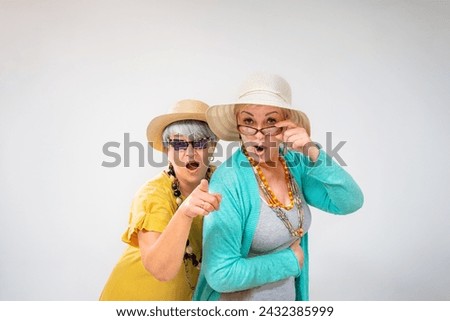 Senior female friends looking at camera with surprise and pointing with finger on gray background