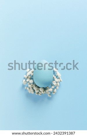 One blue Easter Egg on Bed of White Flowers on sky Blue Background. Easter celebration minimal holiday concept, top view close up pastel colored chicken egg, copy space, vertical monochrome photo