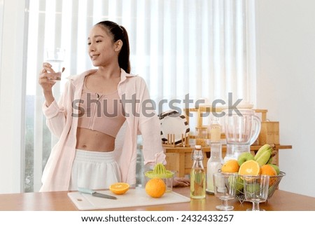 Sporty girl holding a glass of fresh water before drinking in kitchen. Beautiful happy smiling Asian woman preparing healthy smoothie fruit ingredient in blender for making smoothie juice beverage. Royalty-Free Stock Photo #2432433257