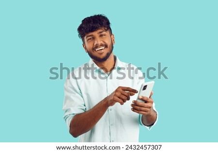 Young happy Indian man holding mobile phone and smiling looking at camera using application for managing business or company dressed in white shirt stands in studio on turquoise background Royalty-Free Stock Photo #2432457307
