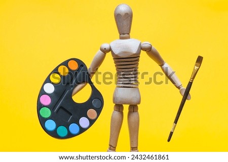 A wooden man holds a palette and a brush in his hand on a yellow background. Artist and creativity concept.