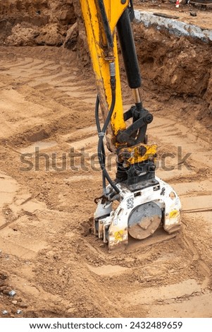 A mini excavator rams the ground with a vibrating plate. Laying of underground sewer pipes and communications during construction. soil compaction. Earthworks, excavation Royalty-Free Stock Photo #2432489659