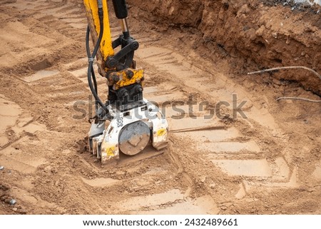 A mini excavator rams the ground with a vibrating plate. Laying of underground sewer pipes and communications during construction. soil compaction. Earthworks, excavation Royalty-Free Stock Photo #2432489661