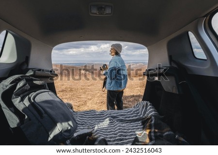 Young happy woman sitting in an open car trunk taking pictures with a camera. Traveling by car, communication in travel concept