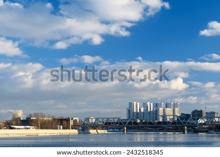 A block of new houses on the river embankment and a railway bridge against the backdrop of a high blue sky with clouds.  Royalty-Free Stock Photo #2432518345