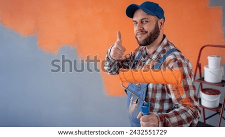 Painter man with beard giving a thumbs up gesture standing in room with painting orange wall. Banner. Copy space