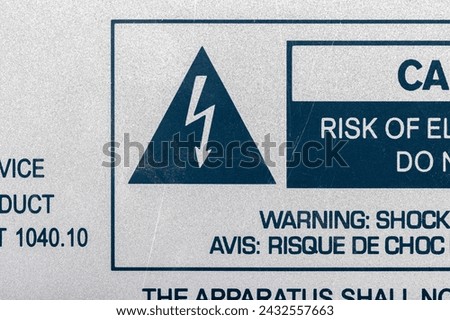A close-up view of a bilingual warning label detailing the risk of electrical shock from a machine device or appliance, with a large lightning bolt symbol inside a triangle, macro detail, nobody