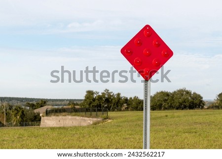 A red traffic sign on a metal pole. There's a grassy meadow and trees in the background. The reflective signage is diamond shaped with embossed round circles. The road end symbol marker is a caution.