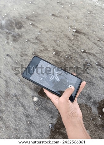 A person holds a smartphone in their hand, capturing a picture of a starfish on the beach. The scene captures the beauty of the starfish against the sandy shore, with the phone held at an angle to get