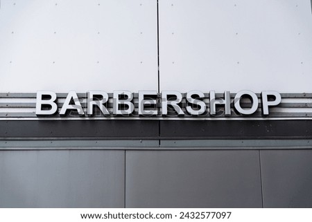 A rectangular grey metal sign with bold font hangs on the facade of the building, serving as the barbershops automotive exterior fixture