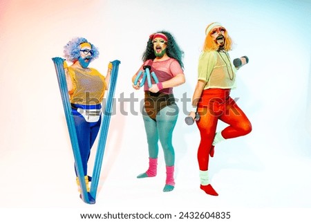 Three drag queens in bright 80s athletic gear enthusiastically performing a workout with weights and elastic band in a studio setting. Royalty-Free Stock Photo #2432604835