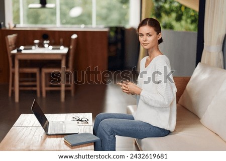 Woman sitting on a cozy modern sofa in her apartment, enjoying a relaxing weekend With a smile on her face, she is comfortably chatting online and shopping for the latest trends, making the most of Royalty-Free Stock Photo #2432619681