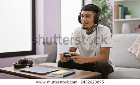 Cheerful young latin man sitting comfortably on a sofa at home, engrossed in playing a gripping video game with wireless control, exuding confidence with a charming smile.