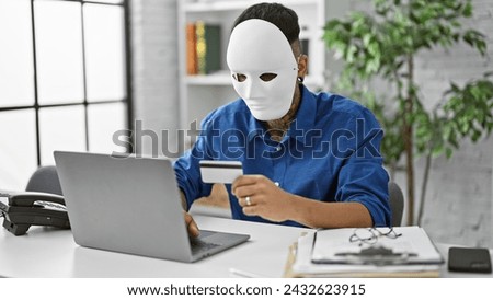 Masked young latin man pulls off a daring credit card hack on his laptop at the office, exposing the dark underbelly of internet crime