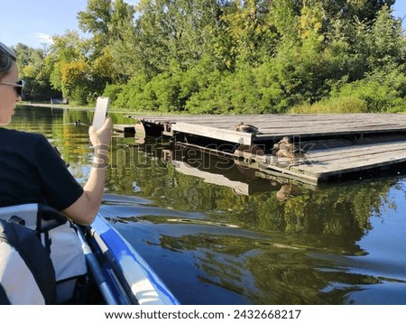 A young woman floats down a river in a boat and takes pictures of wild ducks on a dock on her mobile phone. Active recreation and nature