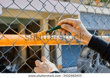 Cropped hand of man painting orange color on chainlink fence Royalty-Free Stock Photo #2432683455