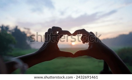 Person making hands love sign against the sun on the field. Hands of woman showing heart shape. Love and compassion concept.
