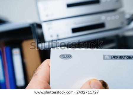 A POV shot features a male hand holding a new SACD package adorned with hi-def jazz signage, with a defocused hi-fi audiophile system in the background, setting the stage for an immersive musical Royalty-Free Stock Photo #2432711727
