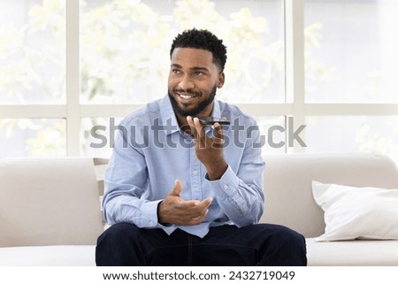 Positive handsome young African business man talking on speaker on cellphone, recording voice message, looking away, thinking, activating modern smart home application
