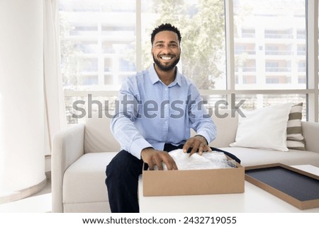 Happy handsome young Black influencer guy unwrapping parcel, opening cardboard package, reviewing purchase from Internet store for shooting, looking at camera, smiling Royalty-Free Stock Photo #2432719055