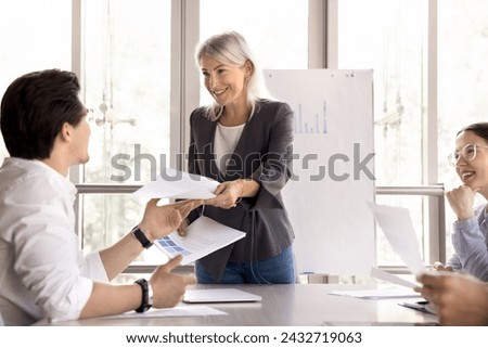 Smiling middle-aged 50s executive businesswoman manager gives handout to colleagues or clients take part in meeting, negotiate in boardroom. Female coach lead new project presentation for investors Royalty-Free Stock Photo #2432719063