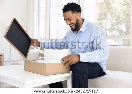 Excited happy young Black consumer man opening cardboard box at home, unpacking awaited purchase from online shop, getting logistic parcel, smiling, laughing in surprise Royalty-Free Stock Photo #2432719117