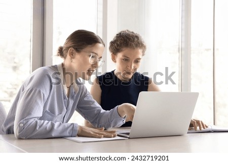 Two young office employees working together use laptop, workmate helps to apprentice with corporate program, new app, software, telling about project, explain task, engaged in teamwork. Apprenticeship Royalty-Free Stock Photo #2432719201