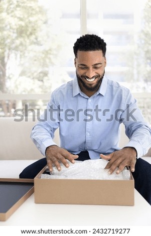 Joyful positive Black client man getting parcel from transportation logistic service, touching plastic wrap in cardboard box, receiving purchase, feeling happy, excited, smiling, laughing Royalty-Free Stock Photo #2432719277