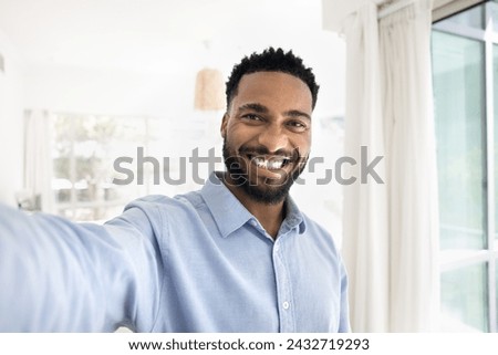 Cheerful handsome young African man taking selfie, looking at camera with toothy smile, showing perfect white teeth, holding gadget with webcam in outstretched hand, talking on video call