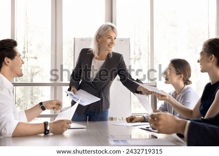 Attractive middle-aged businesslady share handout sheets materials to diverse young colleagues during briefing or seminar event in modern conference room. Paperwork, negotiations meeting in office Royalty-Free Stock Photo #2432719315