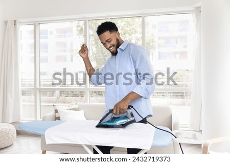 Cheerful young Black man enjoying house work, ironing white fabric towel, dancing to music at board, enjoying domestic activities, laundry, housework at home, having fun