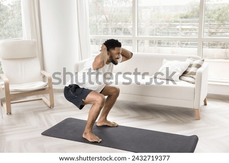 Happy sporty strong young African guy doing morning exercise on yoga mat at home, flexing hips, buttocks muscles in squats, looking away, smiling, enjoying active life style, keeping fit Royalty-Free Stock Photo #2432719377