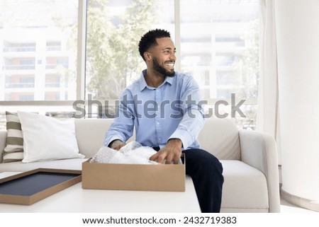 Positive young African American customer guy unpacking parcel at home, opening cardboard box with purchase from online store, looking away with toothy smile, laughing, thinking Royalty-Free Stock Photo #2432719383