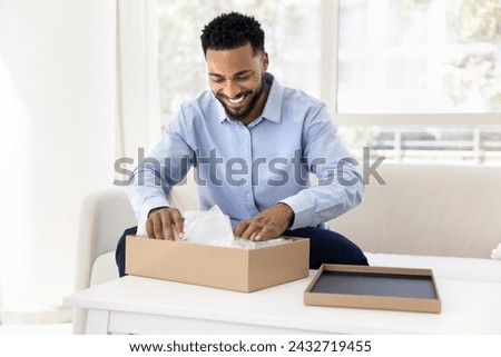 Cheerful young African consumer man getting parcel, opening cardboard box with plastic wrap inside, unwrapping awaited package, cargo from logistic delivery courier service, smiling Royalty-Free Stock Photo #2432719455
