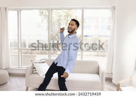 Happy handsome African American guy dancing to music at home, relaxing, celebrating achievement, good news, enjoying activity in white colored apartment interior, having fun, laughing Royalty-Free Stock Photo #2432719459
