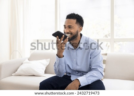 Cheerful young African man dictating voice message on smartphone speaker, using virtual assistant, audio command service, smart home application on mobile phone, talking on speaker call