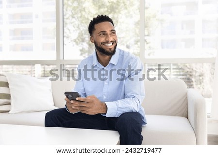Cheerful dreamy African man using gadget for online communication at home, holding mobile phone, looking away, smiling, thinking on media service, application, Internet technology Royalty-Free Stock Photo #2432719477