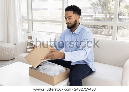 Handsome young African American customer man receiving purchase from Internet store, unpacking logistic cardboard package with plastic wrap, opening carton box at home Royalty-Free Stock Photo #2432719483