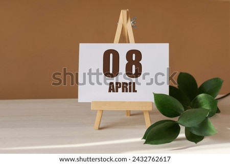 April 8th. Day 8 of month, Calendar date. Green branch, easel with the date and month on desktop. Close-up, brown background. Spring month, day of year concept