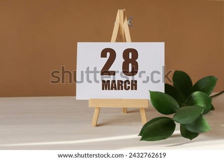 March 28th. Day 28 of month, Calendar date. Green branch, easel with the date and month on desktop. Close-up, brown background. Spring month, day of year concept