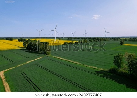 Aerial view Wind turbine on grassy yellow farm canola field against cloudy blue sky in rural area. Offshore windmill park with clouds in farmland Poland Europe. Wind power plant generating electricity Royalty-Free Stock Photo #2432777487