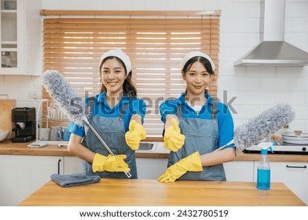 Two Asian cleaning service women on kitchen counter with duster foggy spray and rag illustrate effective housework teamwork. Clean portrait two uniform maid working smiling employee. Royalty-Free Stock Photo #2432780519