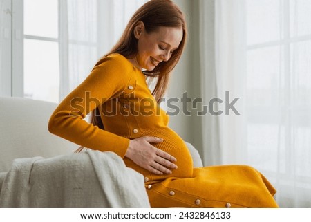 Pregnancy motherhood people expectation future. Pregnant woman with big belly sitting on chair near window at home. Girl hugging her tummy enjoying pregnancy. Maternity tenderness parenthood new life Royalty-Free Stock Photo #2432846133