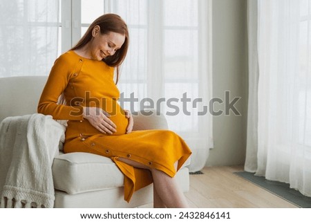 Pregnancy motherhood people expectation future. Pregnant woman with big belly sitting on chair near window at home. Girl hugging her tummy enjoying pregnancy. Maternity tenderness parenthood new life