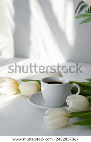 Hot coffee drink cup and white tulip flowers bouquet on table with bright natural sunlight pattern on wall and table, good morning concept, aesthetic business branding template.