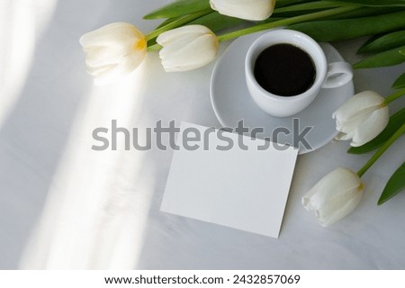 Blank paper card mockup, coffee cup, saucer, white tulip flowers bouquet on neutral marble table background with abstract natural sun light shadows. Good morning concept, postcard, invitation template